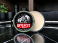 An open round tin of Uppercut Deluxe Matte Pomade sits on a glossy black marble surface, with the cream-colored pomade visible in the clear container and the branded lid leaning in front.