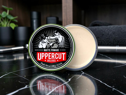 An open round tin of Uppercut Deluxe Matte Pomade sits on a glossy black marble surface, with the cream-colored pomade visible in the clear container and the branded lid leaning in front.