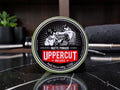A round tin of Uppercut Deluxe Matte Pomade sits on a glossy black marble surface with a blurred bathroom or vanity background.