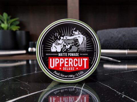 A round tin of Uppercut Deluxe Matte Pomade sits on a glossy black marble surface with a blurred bathroom or vanity background.