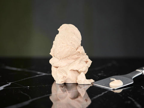 A sculpted beige clay or plaster figure stands on a glossy black marble-like surface, with a metal spatula holding a small dollop of the same beige material beside it.
