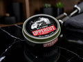 A tin of Uppercut Deluxe Matte Pomade sits on a black marble surface with a blurred grooming setup in the background.
