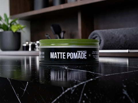 A tin of matte pomade sits on a glossy black marble countertop in a modern bathroom setting, with a blurred towel and sink area in the background.