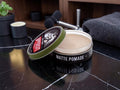 A tin of matte pomade with its lid off, showing the cream-colored pomade inside. The tin has a black label with white text and an olive green rim.