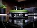 A tin of hair styling product with a black label and a bright green lid, placed on a glossy black marble surface with a blurred bathroom or vanity background.