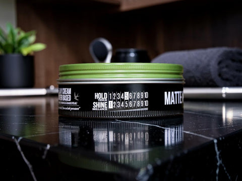 A tin of hair styling product with a black label and a bright green lid, placed on a glossy black marble surface with a blurred bathroom or vanity background.