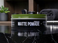 A tin of Brasso Coffee matte pomade sits on a glossy black marble countertop with a blurred bathroom or grooming environment in the background.
