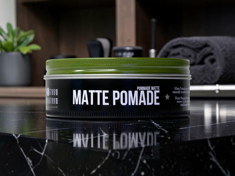A tin of Brasso Coffee matte pomade sits on a glossy black marble countertop with a blurred bathroom or grooming environment in the background.