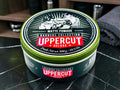 A round tin of Uppercut Deluxe Matte Pomade from the Barbers Collection, placed on a black marble countertop with a folded gray towel and barbershop accessories blurred in the background.