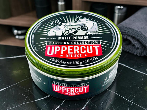 A round tin of Uppercut Deluxe Matte Pomade from the Barbers Collection, placed on a black marble countertop with a folded gray towel and barbershop accessories blurred in the background.