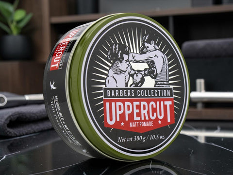 A 300g tin of Uppercut Matt Pomade from the Barbers Collection, featuring an olive green and black container with a vintage-style circular label, set on a dark reflective surface.