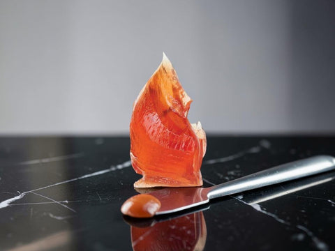 A small sculpted piece of translucent orange-red wax melt or resin standing upright on a glossy black marble surface, with a metal spatula and a small droplet of the same material in front.