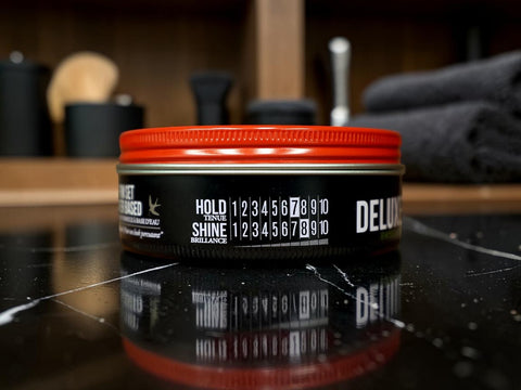 A tin of Menage au Chevelu Shine Delux hair product with a bright red lid and black label, set on a reflective black marble surface with a blurred bathroom background.
