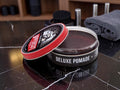An open tin of L'Envers Deluxe Pomade with its lid leaning against the container, set on a dark marble surface with grooming accessories blurred in the background.