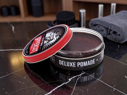An open tin of L'Envers Deluxe Pomade with its lid leaning against the container, set on a dark marble surface with grooming accessories blurred in the background.