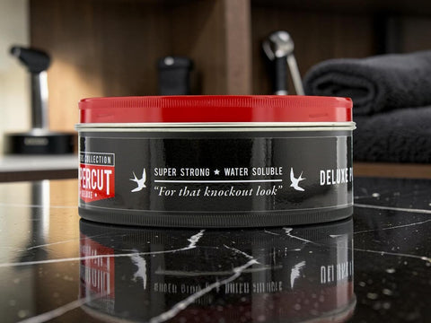 A tin of Percut Deluxe Hair Styling Product, featuring a black body and a bright red lid, set on a reflective black marble surface with a blurred salon background.