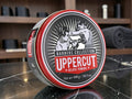 A round tin of UpperCut Barbers Collection Deluxe Pomade sits on a dark marble countertop with a blurred barbershop background.