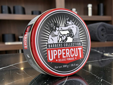A round tin of UpperCut Barbers Collection Deluxe Pomade sits on a dark marble countertop with a blurred barbershop background.
