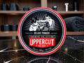 A round tin of Uppercut Deluxe Pomade from the Barbers Collection, displayed on a dark marble countertop with a blurred barbershop background.