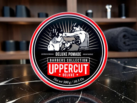 A round tin of Uppercut Deluxe Pomade from the Barbers Collection, displayed on a dark marble countertop with a blurred barbershop background.