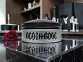 A cosmetic jar labeled 'REGEN-ROSE' sits on a glossy black marble surface with a red rose beside it and a softly blurred bathroom or vanity background.