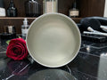 A round, matte off-white cosmetic bowl or compact tin shown from a top-down view on a black marble surface, with a red rose and skincare bottles in the background.