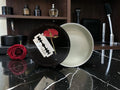 A circular black cosmetic container with a matching lid leaning against it on a glossy black marble surface. The lid features a red rose and a white razor graphic, while the open container shows a pale cream interior.
