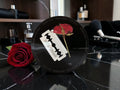A circular black collectible plaque featuring a red rose and a white razor blade graphic, styled on a black marble surface next to a real red rose with perfume bottles in the background.