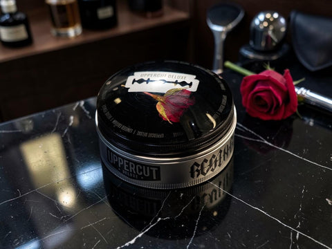 A black metal tin of Uppercut Regime grooming product sits on a glossy black marble surface, with a red rose and grooming tools blurred in the background.