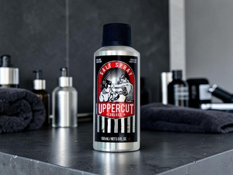 A 500 mL can of Uppercut Deluxe Hair Spray standing on a dark countertop, with rolled towels and other grooming bottles blurred in the background.