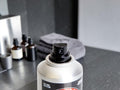 A close-up of a silver spray can with a black pump dispenser, set on a dark countertop with skincare bottles and a gray towel blurred in the background.