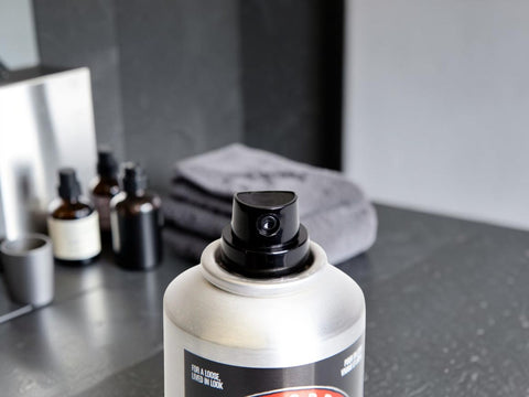 A close-up of a silver spray can with a black pump dispenser, set on a dark countertop with skincare bottles and a gray towel blurred in the background.