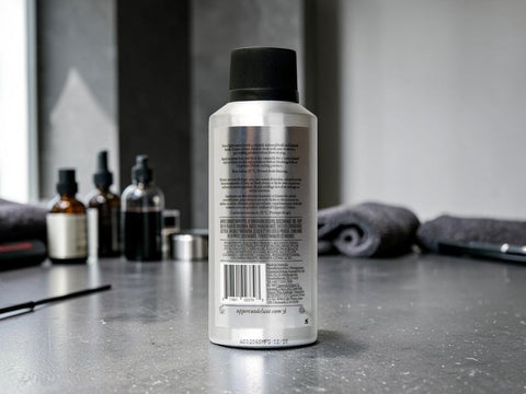 A silver cylindrical bottle with a black cap stands on a gray countertop, showing the back label. The background is a blurred bathroom or grooming setting with dark towels and other bottles.