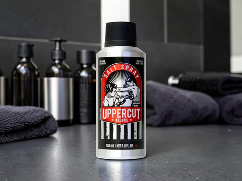 A bottle of Uppercut Deluxe Salt Spray standing on a dark countertop, with folded towels and dark bottles blurred in the background.