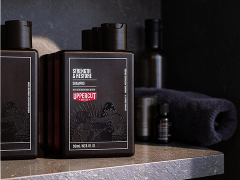 A dark brown rectangular bottle of Uppercut & Regain Shampoo on a stone countertop, with a matching towel and other grooming products blurred in the background.