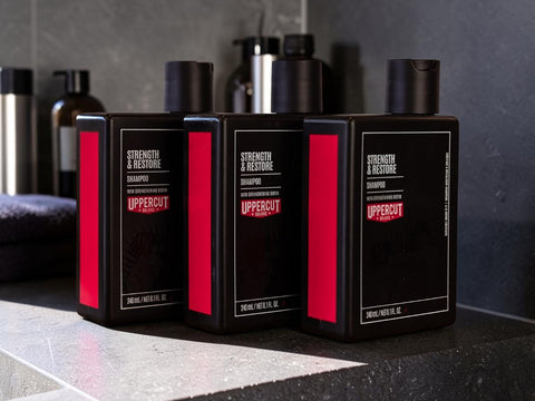 Three black Uppercut Hair Strength & Restore shampoo bottles with red side panels and black caps, arranged on a dark countertop in a bathroom setting.