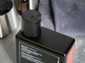 A close-up of a black rectangular bottle with a matching black flip-top cap, photographed on a countertop with a gray towel and metallic container in the background.
