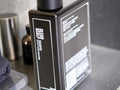 A black rectangular bottle of Clear Shampoo with white text, shown on a gray countertop with a dark towel and other bathroom items blurred in the background.