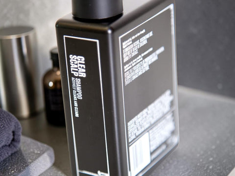 A black rectangular bottle of Clear Shampoo with white text, shown on a gray countertop with a dark towel and other bathroom items blurred in the background.