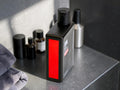 A black rectangular bottle with a bold red front panel and black cap, standing on a dark countertop beside folded gray towels and other toiletries.
