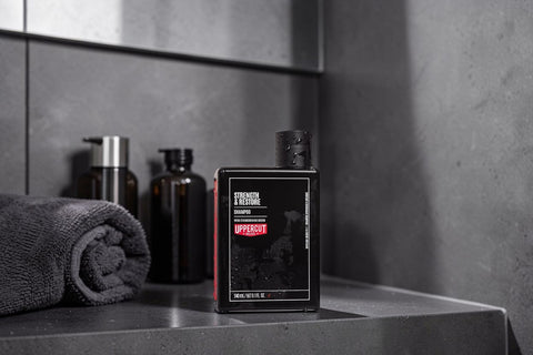 A black rectangular bottle of UpperCrest Strength & Restore haircare product standing on a dark countertop, with a rolled towel and dark bottles blurred in the background against a gray tiled wall.