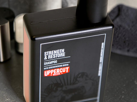 A close-up of an UpperCut Strength & Restore Shampoo bottle, showing its matte black packaging with a white and red label, set against a dark, blurred background.