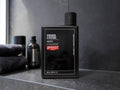 A black rectangular bottle of Uppercut Strength & Restore Shampoo stands on a dark countertop in a bathroom setting, with folded towels and another bottle blurred in the background.