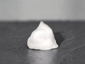 A small white, dome-shaped object with a smooth, matte surface sits centered on a dark, textured surface against a neutral gray background.