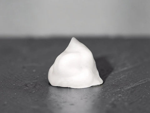 A small white, dome-shaped object with a smooth, matte surface sits centered on a dark, textured surface against a neutral gray background.