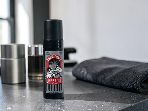 A bottle of Uppercut Foam Torch hair product stands on a dark countertop, with a folded dark towel and metallic containers blurred in the background.