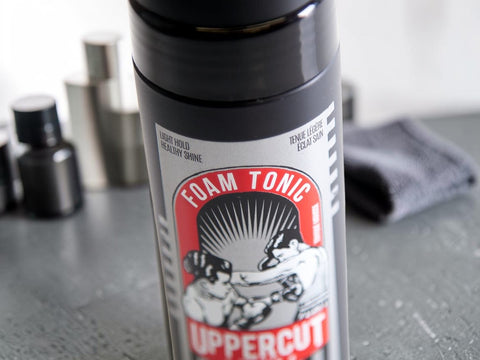 A close-up of a black cylindrical foam tonic spray bottle labeled 'FOAM TONIC UPPERCUT' with a red, white, and black graphic, standing on a gray surface with blurred bottles and a cloth in the background.