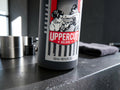 A close-up of a dark gray Uppercut Deluxe 150 mL bottle on a dark countertop, with a blurred gym or bathroom background including a towel and metal containers.