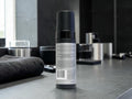 A matte black cylindrical bottle with a white label stands on a dark countertop in a modern bathroom setting with a towel and metallic containers blurred in the background.