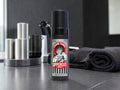 A bottle of Uppercut Team Tonic hair product standing on a dark countertop, with a blurred modern grooming setup in the background including metallic containers and a folded dark towel.